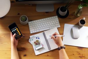 Vista dall'alto di una scrivania in legno con un computer, tastiera, tazza di caffè e piante, dove una persona calcola cifre su uno smartphone, prende appunti su un quaderno e ha banconote e monete in euro accanto.