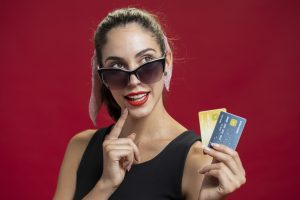 Una giovane donna con occhiali da sole eleganti e rossetto rosso sorride mentre tiene due carte di credito colorate in mano, indossando un vestito nero e una fascia rosa tra i capelli, davanti a uno sfondo rosso.