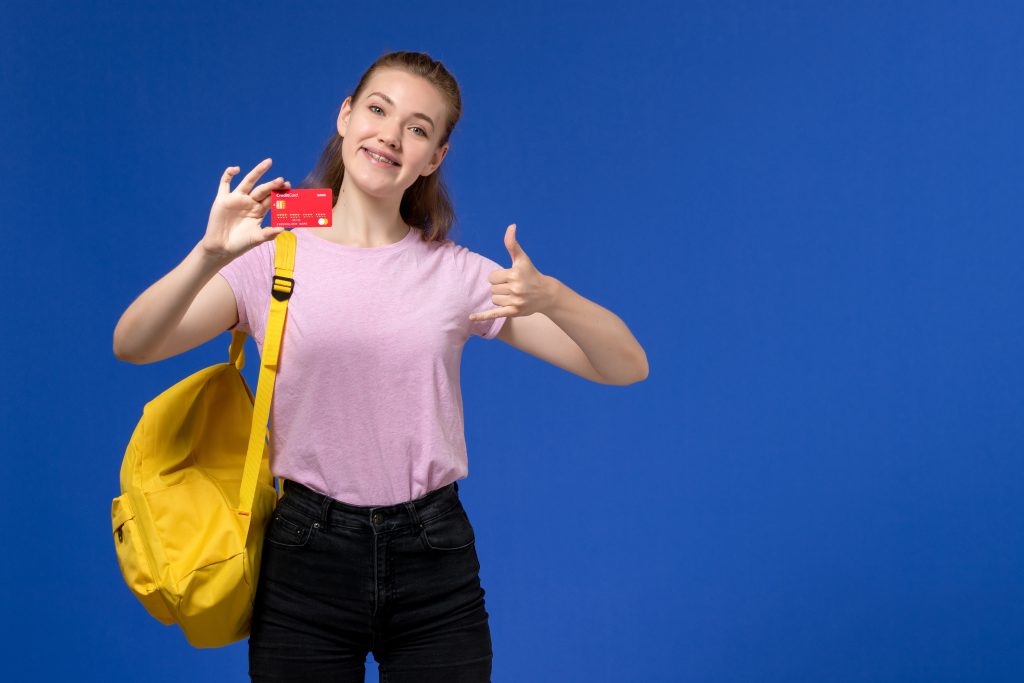 Giovane ragazza con zaino giallo e maglietta rosa sorride mostrando una carta di credito rossa, su sfondo blu, simboleggiando indipendenza finanziaria e gestione responsabile del denaro.