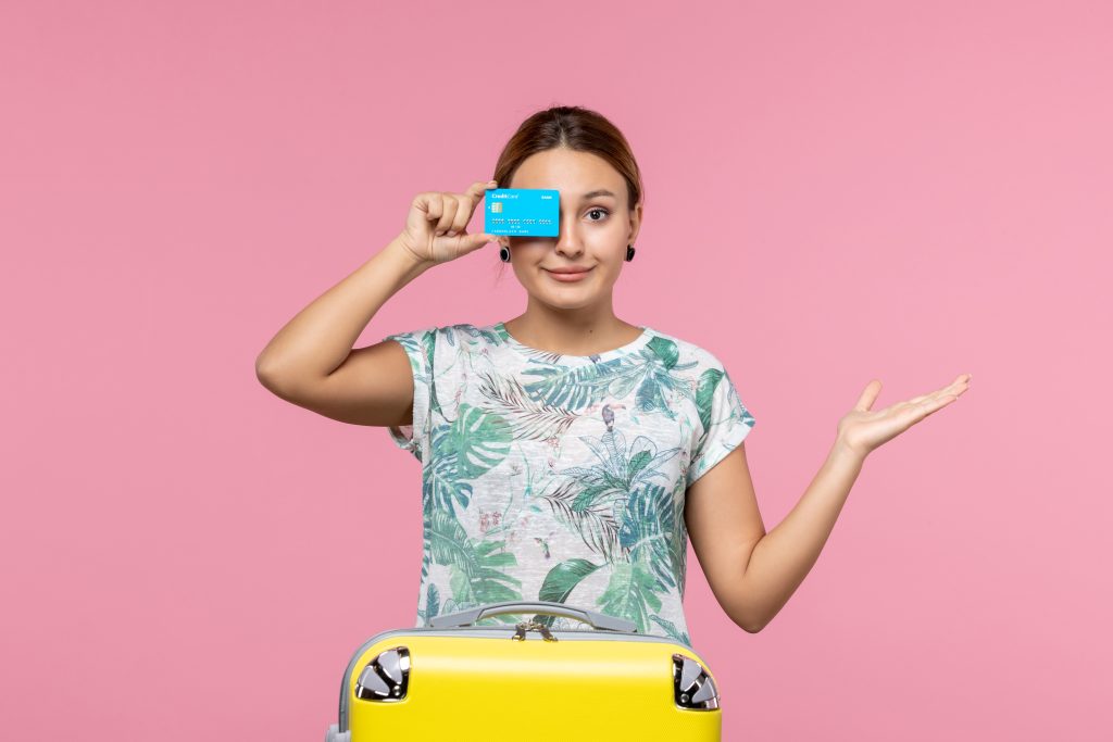 Giovane donna con maglietta tropicale tiene una carta di credito blu davanti all'occhio e una valigia gialla davanti a sé, su sfondo rosa, simboleggiando viaggi e spese con carta.