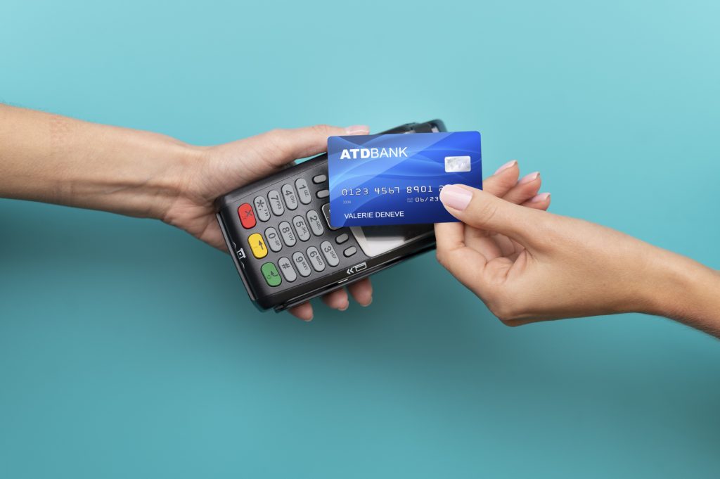 Una persona sta effettuando un pagamento senza contatto con una carta di credito blu di ATD Bank, avvicinandola a un terminale POS tenuto da un'altra persona. Lo sfondo azzurro crea un contrasto moderno e minimalista, evidenziando la semplicità e la rapidità delle transazioni digitali nei pagamenti quotidiani.