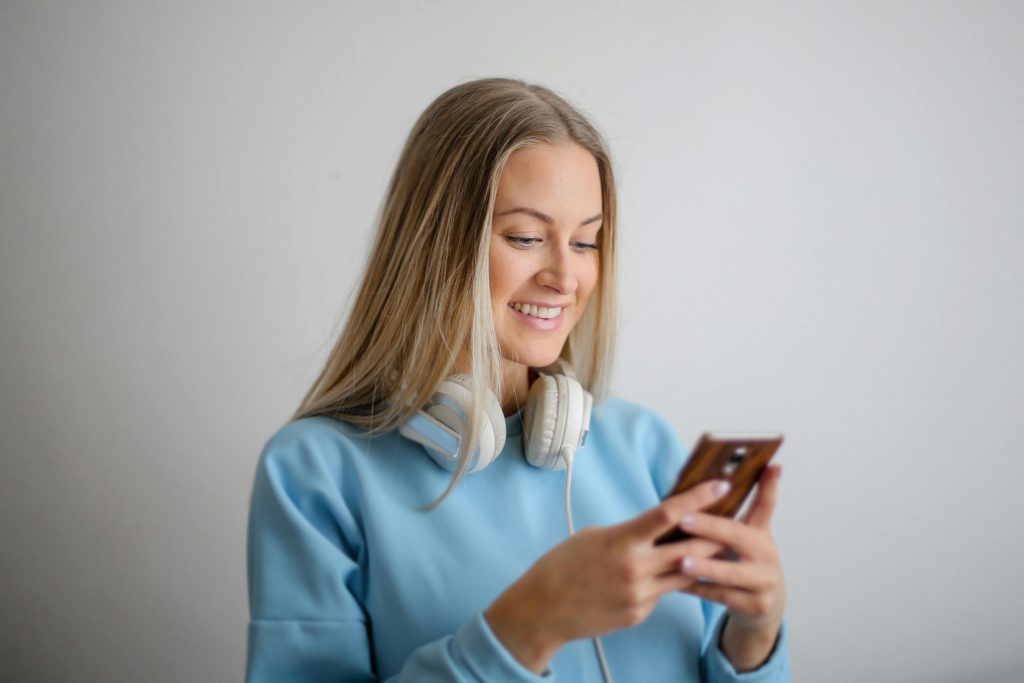 Una giovane donna con lunghi capelli biondi indossa una felpa azzurra e ha un paio di cuffie bianche intorno al collo. Sorride mentre guarda il suo smartphone, apparentemente immersa in una conversazione o nella navigazione online. L'immagine trasmette un senso di relax e connessione digitale.