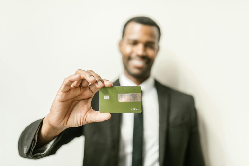 A smiling man in a black suit and tie holds a green credit card towards the camera, with a blurred background. The image conveys confidence and financial empowerment, symbolizing banking, credit services, or financial success.