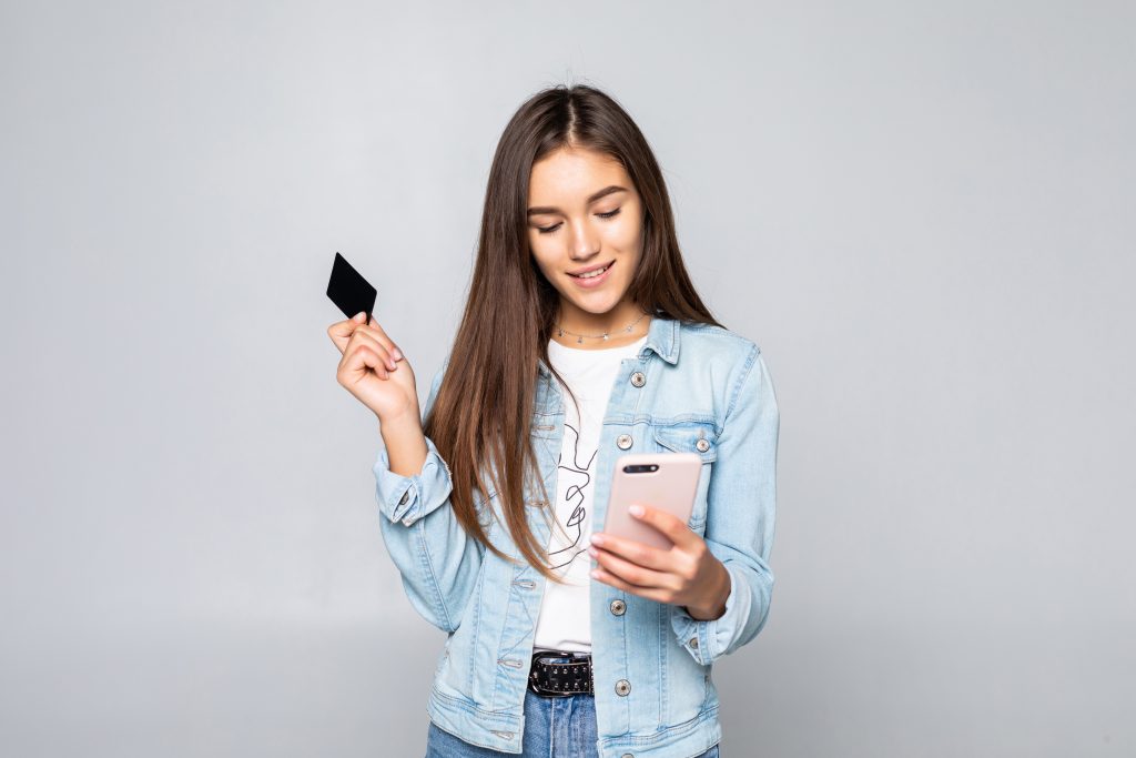 Una giovane donna con i capelli lunghi e una giacca di jeans sorride mentre tiene una carta di credito nera in una mano e uno smartphone nell'altra. L'espressione felice suggerisce che stia effettuando un acquisto online o gestendo una transazione digitale. Lo sfondo neutro evidenzia il concetto di shopping moderno e pagamenti digitali.