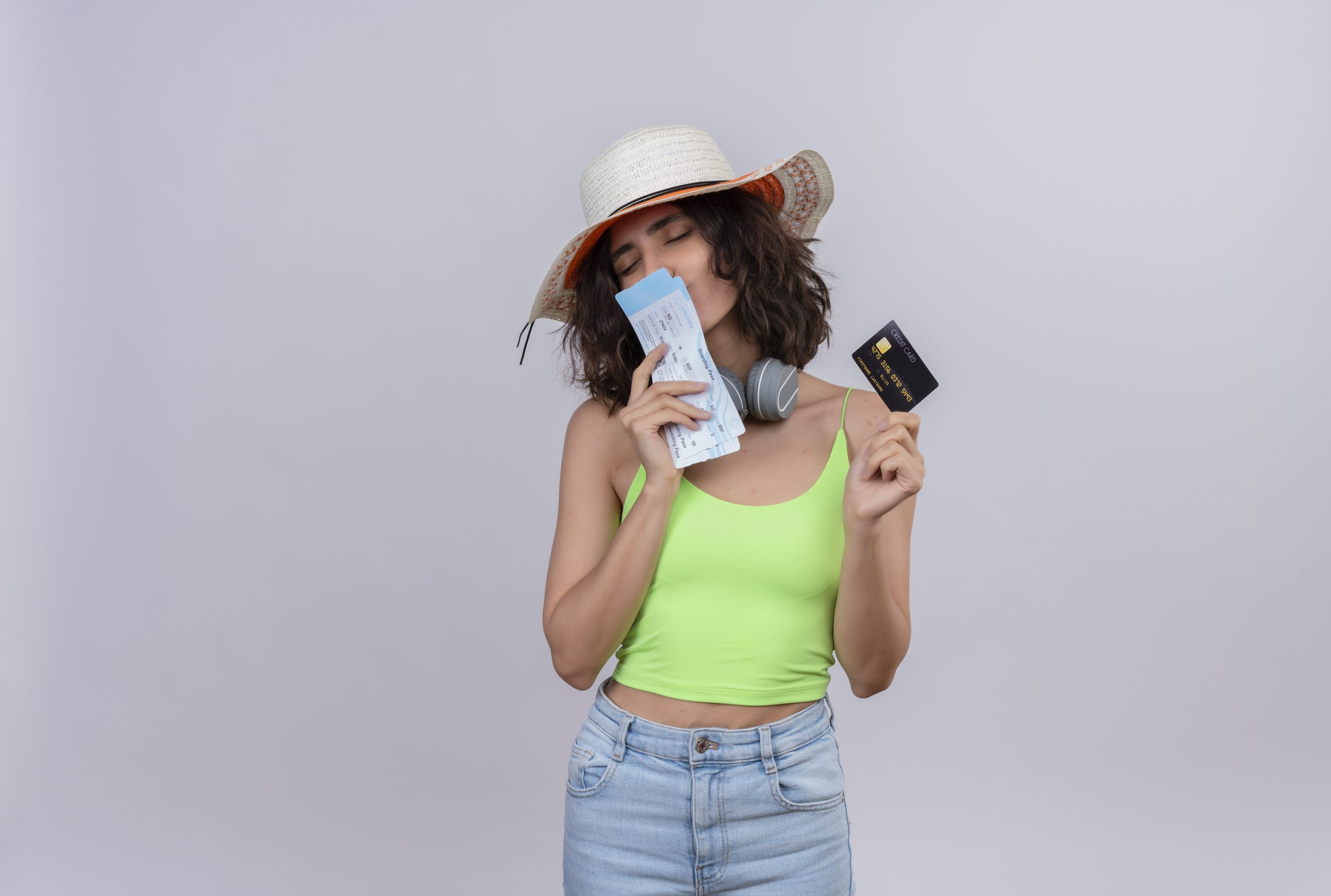 Una giovane donna con cappello da sole e cuffie al collo sorride con gli occhi chiusi mentre bacia dei biglietti aerei e tiene una carta di credito in mano, indossando un top verde e jeans chiari su uno sfondo bianco.