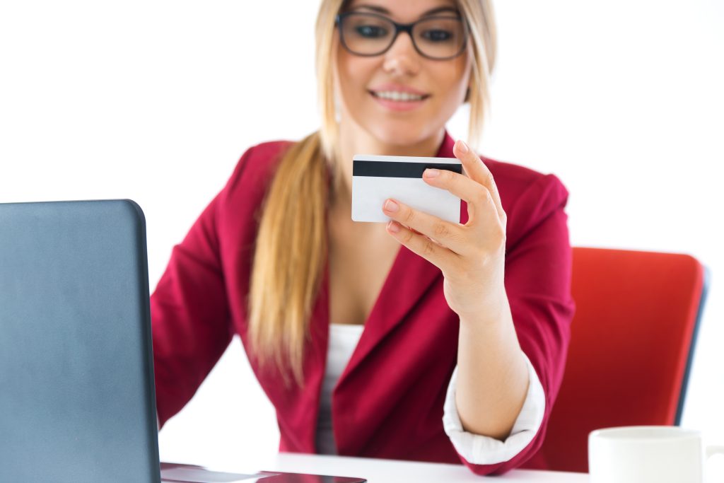 Una giovane donna con gli occhiali e una giacca rossa sorride mentre tiene in mano una carta di credito, seduta alla scrivania con un laptop davanti. La scena rappresenta lo shopping online, le transazioni digitali o l'uso delle carte di pagamento nel mondo degli affari moderni.