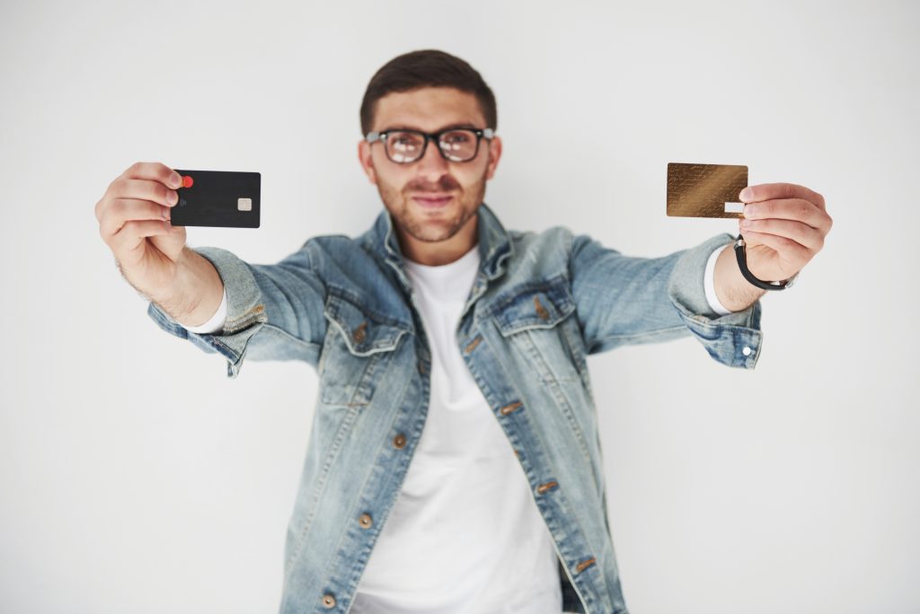 Un giovane uomo con occhiali e giacca di jeans sorride mentre tiene in mano due carte di credito, una nera e una dorata, mostrandole verso la fotocamera su uno sfondo bianco.
