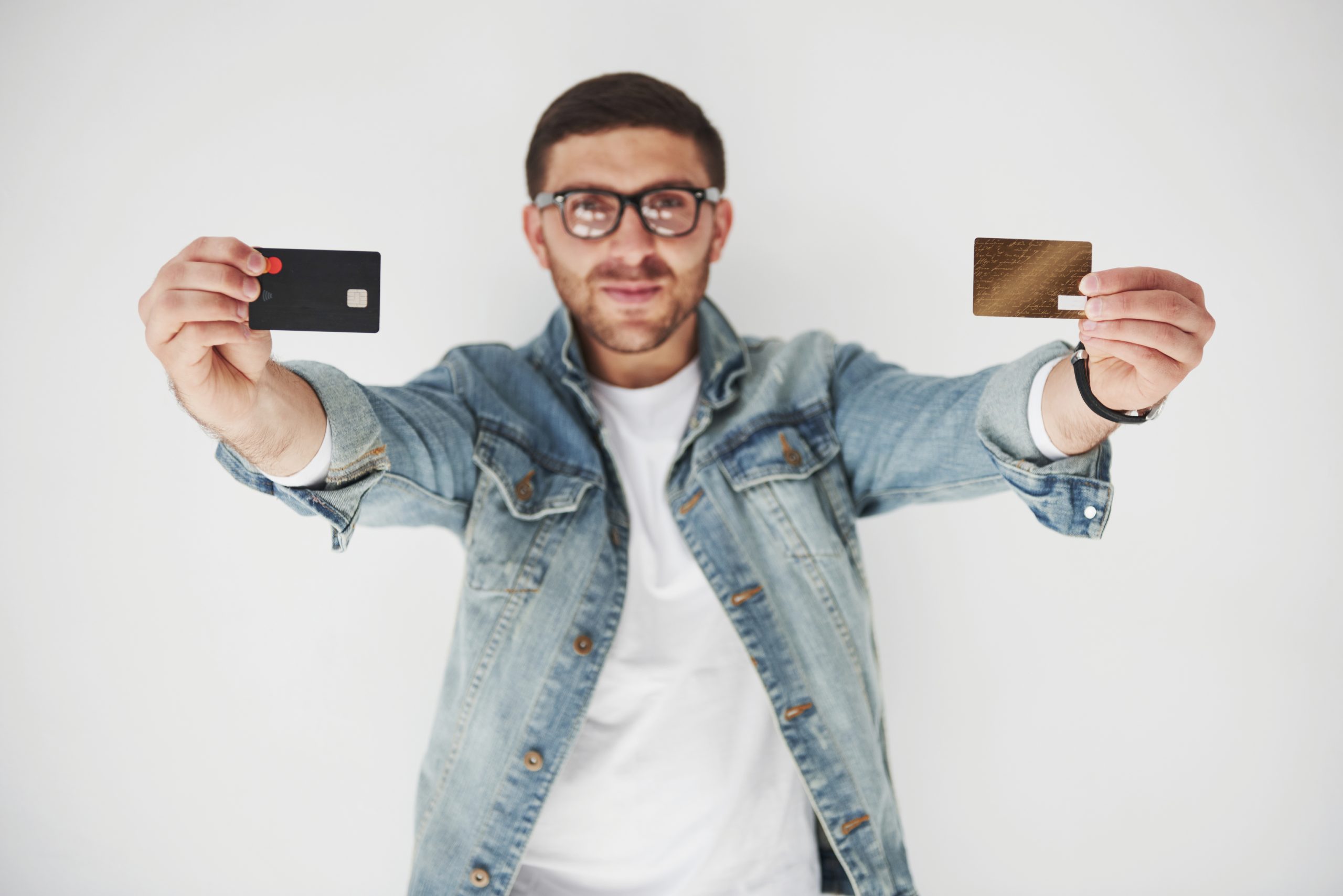 Un giovane uomo con occhiali e giacca di jeans sorride mentre tiene in mano due carte di credito, una nera e una dorata, mostrandole verso la fotocamera su uno sfondo bianco.