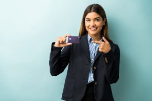 Giovane donna d'affari sorridente in abito formale, in piedi davanti a uno sfondo azzurro mentre mostra e indica una carta di credito viola. L'immagine rappresenta fiducia e professionalità nel contesto delle carte di credito aziendali, evidenziando strumenti finanziari pensati per facilitare la gestione delle spese aziendali in modo sicuro ed efficiente.