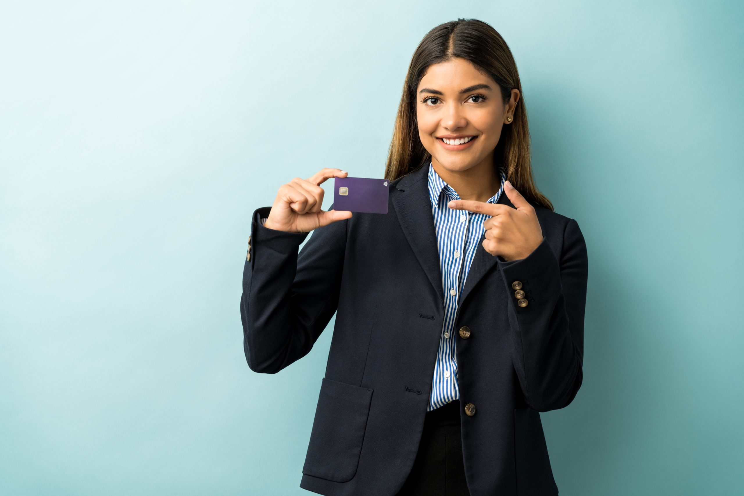 Giovane donna d'affari sorridente in abito formale, in piedi davanti a uno sfondo azzurro mentre mostra e indica una carta di credito viola. L'immagine rappresenta fiducia e professionalità nel contesto delle carte di credito aziendali, evidenziando strumenti finanziari pensati per facilitare la gestione delle spese aziendali in modo sicuro ed efficiente.