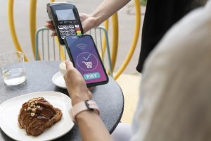 Una persona sta effettuando un pagamento contactless con lo smartphone in un bar all’aperto. Sul tavolo ci sono un bicchiere d’acqua, una tazza di cappuccino e un cornetto con nocciole. Un’altra persona tiene il lettore di carte, mentre lo schermo del telefono mostra l’icona di pagamento completato.