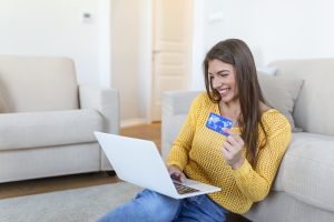Giovane donna sorridente seduta sul pavimento del soggiorno mentre fa shopping online con un laptop, tenendo in mano una carta di credito blu.