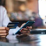 Donna seduta in un bar con una camicia bianca utilizza uno smartphone e tiene una carta di credito in mano, mentre beve un caffè da asporto; scena rappresentativa dell’impatto delle regolamentazioni europee sui metodi di pagamento digitali e sull’e-commerce quotidiano.