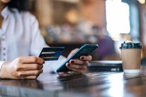 Donna seduta in un bar con una camicia bianca utilizza uno smartphone e tiene una carta di credito in mano, mentre beve un caffè da asporto; scena rappresentativa dell’impatto delle regolamentazioni europee sui metodi di pagamento digitali e sull’e-commerce quotidiano.