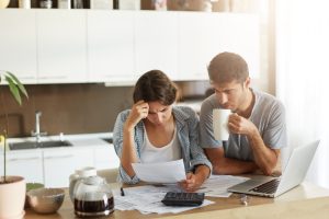 Una giovane coppia seduta in cucina guarda preoccupata dei documenti finanziari. La donna tiene una mano sulla fronte mentre legge una bolletta, e l’uomo, con una tazza in mano, osserva i fogli. Sul tavolo ci sono una calcolatrice, una caffettiera, alcune tazze e un computer portatile, suggerendo che stanno cercando di gestire il loro bilancio familiare.