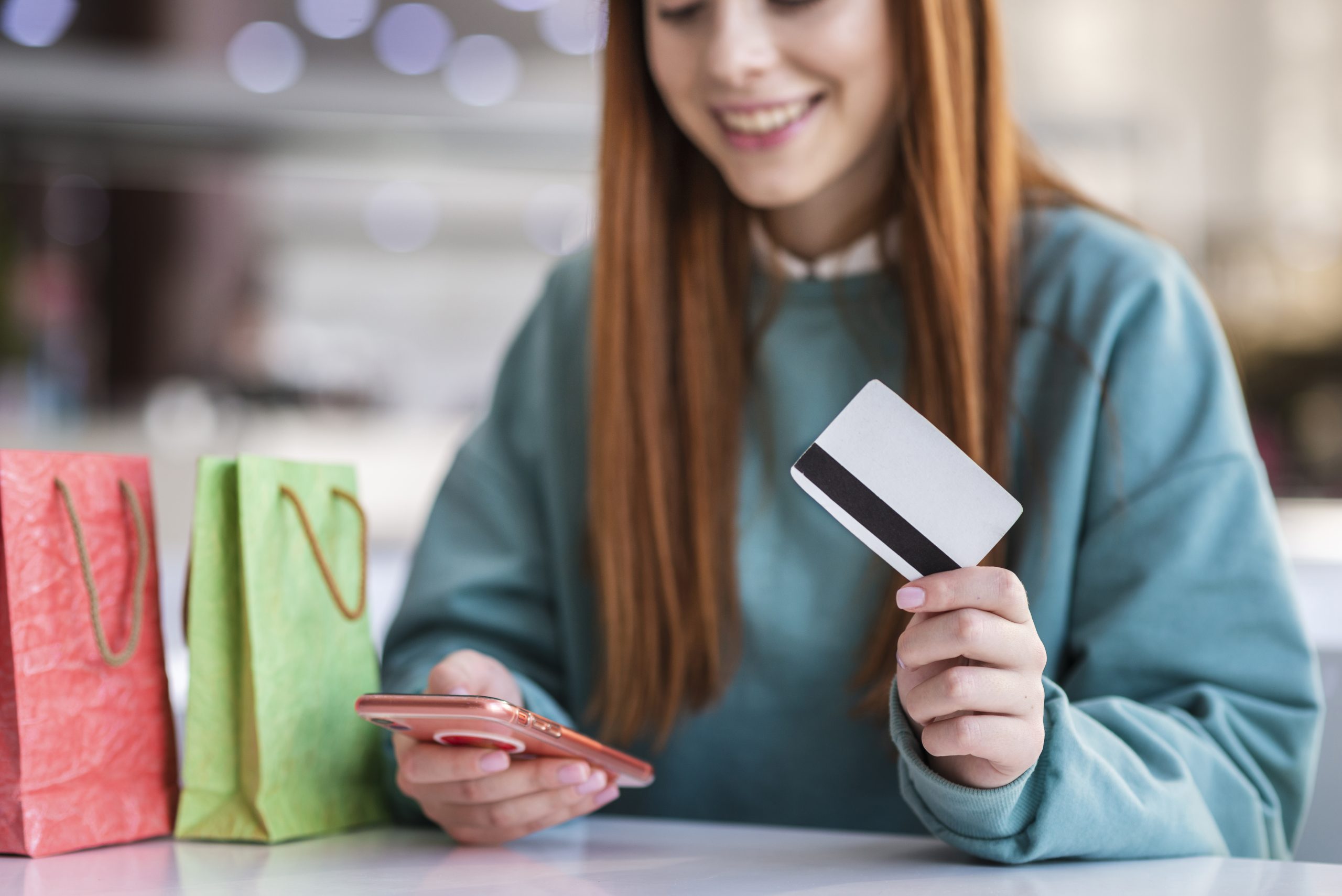 Giovane donna sorride mentre effettua un acquisto online utilizzando il suo smartphone e una carta di credito. Sul tavolo ci sono delle borse colorate da shopping. L’immagine rappresenta un’esperienza d’acquisto moderna e positiva, ideale per illustrare l’uso di carte di credito con cashback.