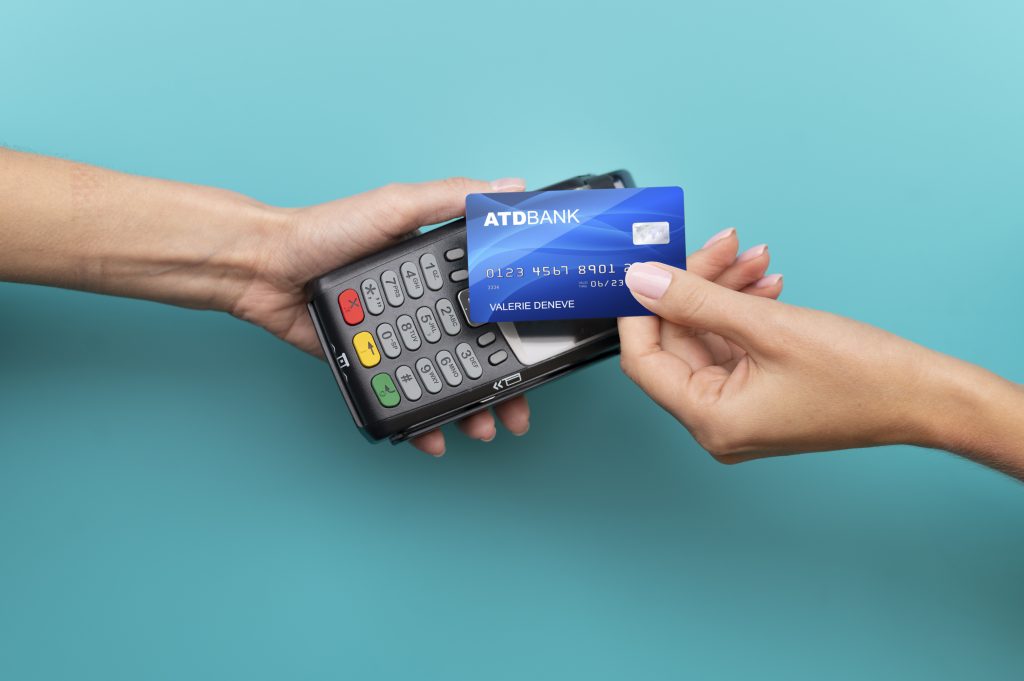Una persona effettua un pagamento utilizzando una delle moderne carte di credito contactless, avvicinando la propria carta blu con il logo "ATDBANK" a un terminale POS. Un'altra mano tiene il lettore di carte, pronto a ricevere la transazione senza bisogno di inserire il chip o digitare il PIN. Lo sfondo è di colore turchese, creando un contrasto vivido con i dispositivi elettronici e le mani.