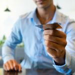 Un uomo in camicia azzurra, seduto a un tavolo in un ambiente moderno e luminoso, porge una carta di credito con la mano destra. L'inquadratura è focalizzata sulla carta, mentre il volto dell'uomo è sfocato. L'immagine rappresenta un gesto quotidiano legato al pagamento digitale e sottolinea l'importanza delle carte di credito come strumento essenziale per la gestione delle spese personali e professionali, in particolare per lavoratori autonomi e freelance.