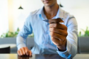 Un uomo in camicia azzurra, seduto a un tavolo in un ambiente moderno e luminoso, porge una carta di credito con la mano destra. L'inquadratura è focalizzata sulla carta, mentre il volto dell'uomo è sfocato. L'immagine rappresenta un gesto quotidiano legato al pagamento digitale e sottolinea l'importanza delle carte di credito come strumento essenziale per la gestione delle spese personali e professionali, in particolare per lavoratori autonomi e freelance.