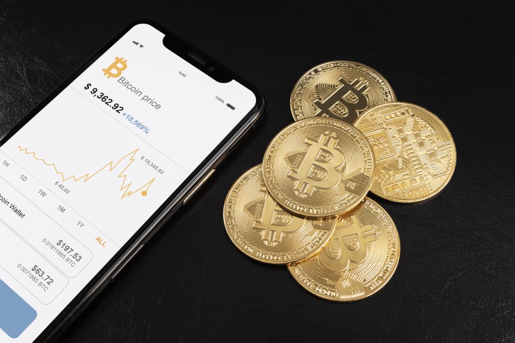 Uno smartphone mostra il prezzo del Bitcoin e un grafico di andamento, affiancato da alcune monete fisiche dorate con il simbolo del Bitcoin, il tutto su sfondo nero. L'immagine rappresenta l'uso crescente delle tecnologie mobili per monitorare gli investimenti in criptovalute. Questo contesto è particolarmente rilevante per l'espansione delle carte premio per criptovalute in Italia, che permettono agli utenti di convertire punti fedeltà in asset digitali.