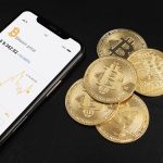 Uno smartphone mostra il prezzo del Bitcoin e un grafico di andamento, affiancato da alcune monete fisiche dorate con il simbolo del Bitcoin, il tutto su sfondo nero. L'immagine rappresenta l'uso crescente delle tecnologie mobili per monitorare gli investimenti in criptovalute. Questo contesto è particolarmente rilevante per l'espansione delle carte premio per criptovalute in Italia, che permettono agli utenti di convertire punti fedeltà in asset digitali.
