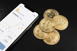 Uno smartphone mostra il prezzo del Bitcoin e un grafico di andamento, affiancato da alcune monete fisiche dorate con il simbolo del Bitcoin, il tutto su sfondo nero. L'immagine rappresenta l'uso crescente delle tecnologie mobili per monitorare gli investimenti in criptovalute. Questo contesto è particolarmente rilevante per l'espansione delle carte premio per criptovalute in Italia, che permettono agli utenti di convertire punti fedeltà in asset digitali.