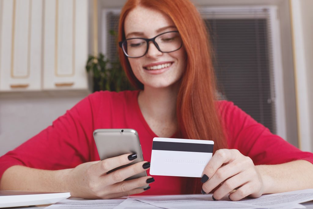 Una giovane donna dai capelli rossi sorride mentre utilizza il suo smartphone e tiene in mano una carta di credito. È seduta a un tavolo con documenti, probabilmente gestendo pagamenti o acquisti online. L'immagine rappresenta un uso comune della carta di credito in Italia per operazioni digitali quotidiane.