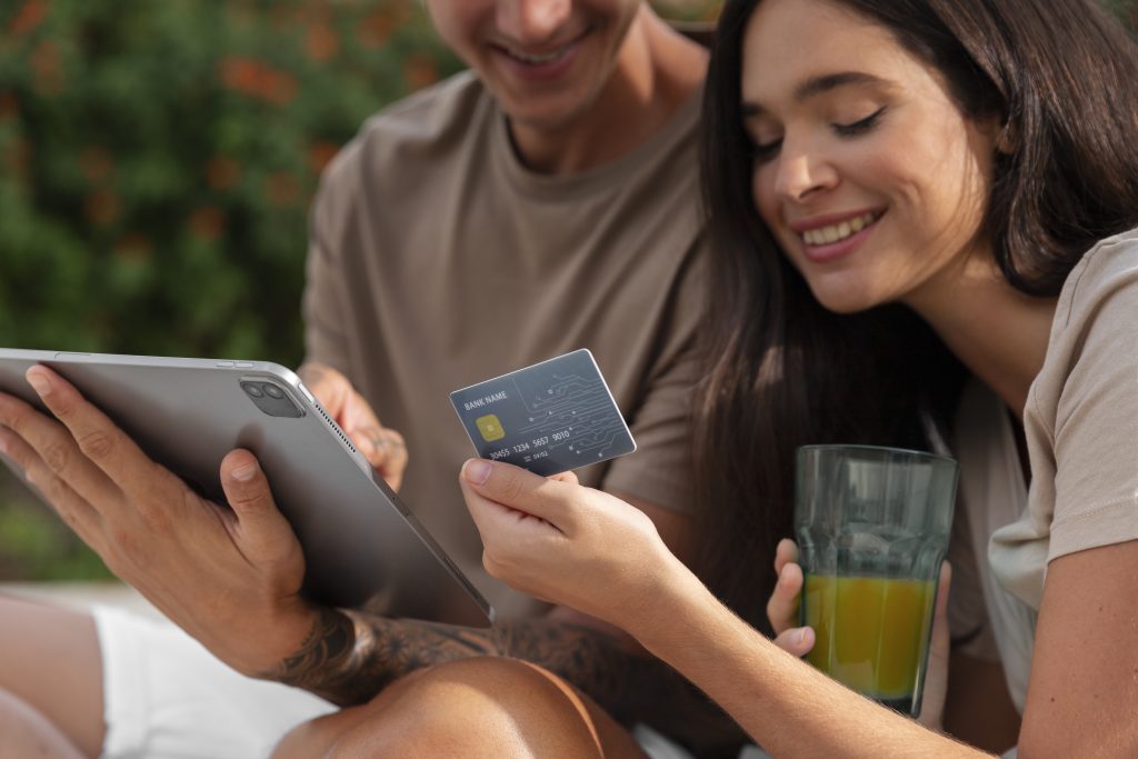 Una giovane coppia è seduta all’aperto mentre sorride e consulta un tablet. La donna tiene in mano una carta di credito con design tecnologico, mentre nell’altra mano ha un bicchiere con succo d’arancia. L’atmosfera è rilassata e familiare, suggerendo l’uso condiviso e sicuro di una carta di credito per famiglie pensata per la gestione quotidiana delle spese.