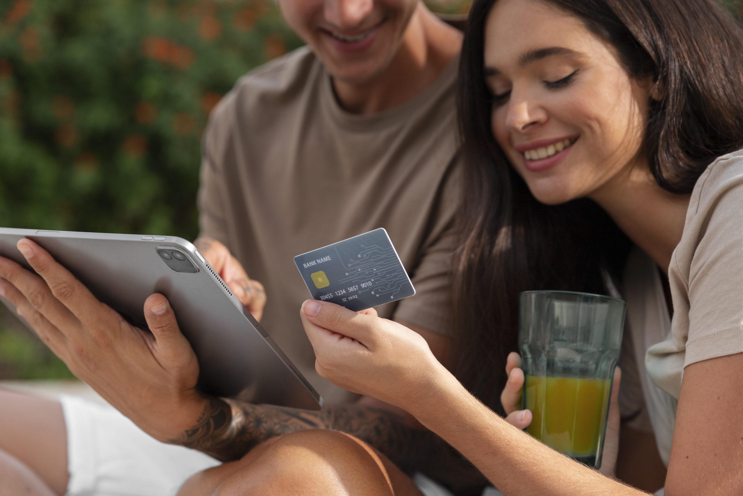 Una giovane coppia è seduta all’aperto mentre sorride e consulta un tablet. La donna tiene in mano una carta di credito con design tecnologico, mentre nell’altra mano ha un bicchiere con succo d’arancia. L’atmosfera è rilassata e familiare, suggerendo l’uso condiviso e sicuro di una carta di credito per famiglie pensata per la gestione quotidiana delle spese.