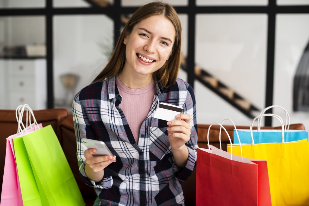 Una giovane donna sorridente è seduta su un divano con diverse borse colorate per lo shopping accanto a sé. Tiene in una mano uno smartphone e nell’altra una delle sue carte di credito, suggerendo che sta effettuando acquisti online o controllando i suoi pagamenti digitali.
