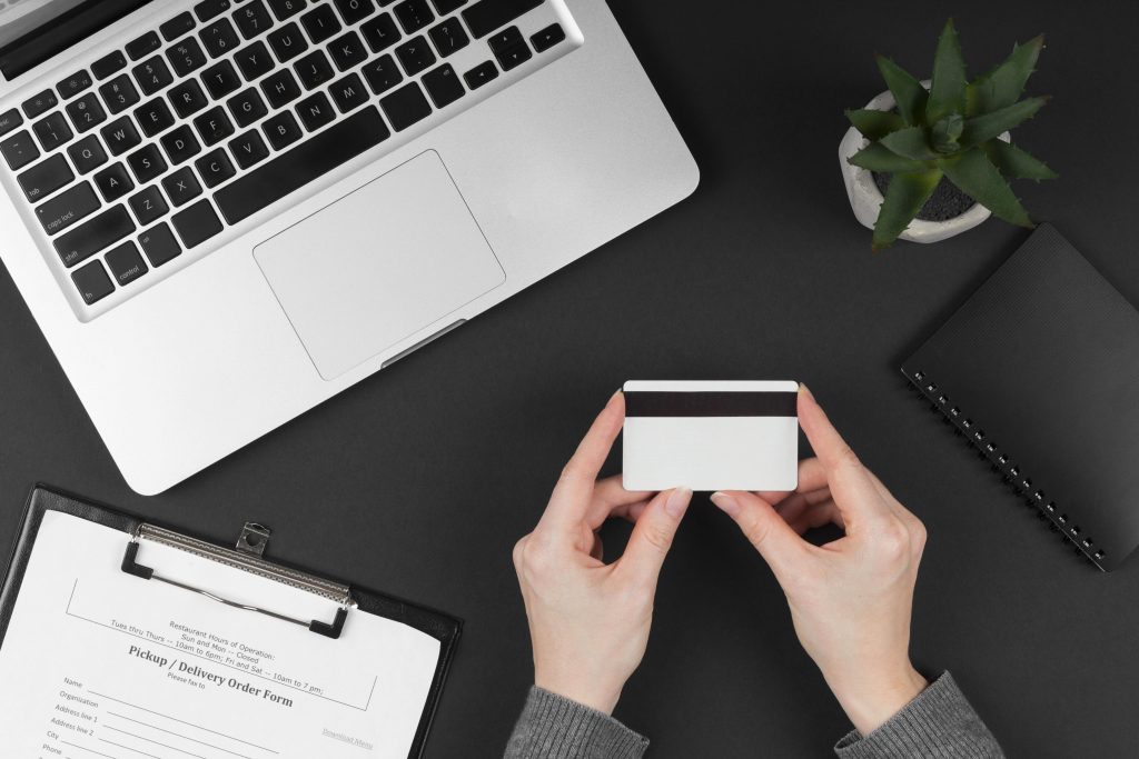Vista dall’alto di una scrivania moderna con un laptop, una pianta decorativa, un taccuino nero e un modulo per ordini di consegna. Al centro, una persona tiene in mano una carta di credito. L’immagine evoca un ambiente professionale e organizzato, perfetto per rappresentare l’uso di carte di credito per le piccole imprese.