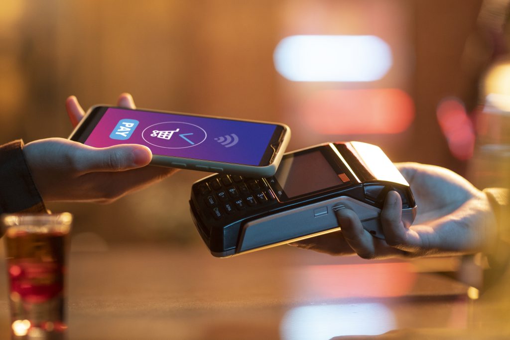 Un telefono cellulare viene avvicinato a un terminale di pagamento POS per completare una transazione contactless tramite un'app di pagamento digitale. Lo sfondo sfocato con luci calde suggerisce un ambiente moderno e urbano. Questa scena rappresenta l’uso crescente dei credito virtuali in Italia, che consente pagamenti rapidi e sicuri senza l’uso di carte fisiche.