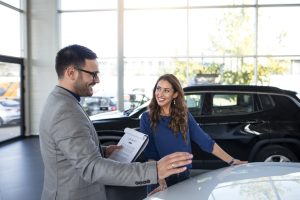 Una donna sorridente discute con un venditore all'interno di una concessionaria moderna e luminosa. Sullo sfondo, si intravedono diverse auto parcheggiate, mentre l'uomo, elegante con un completo grigio, tiene in mano un contratto. La scena rappresenta un momento positivo e cordiale, tipico del processo di finanziamento auto in Italia.