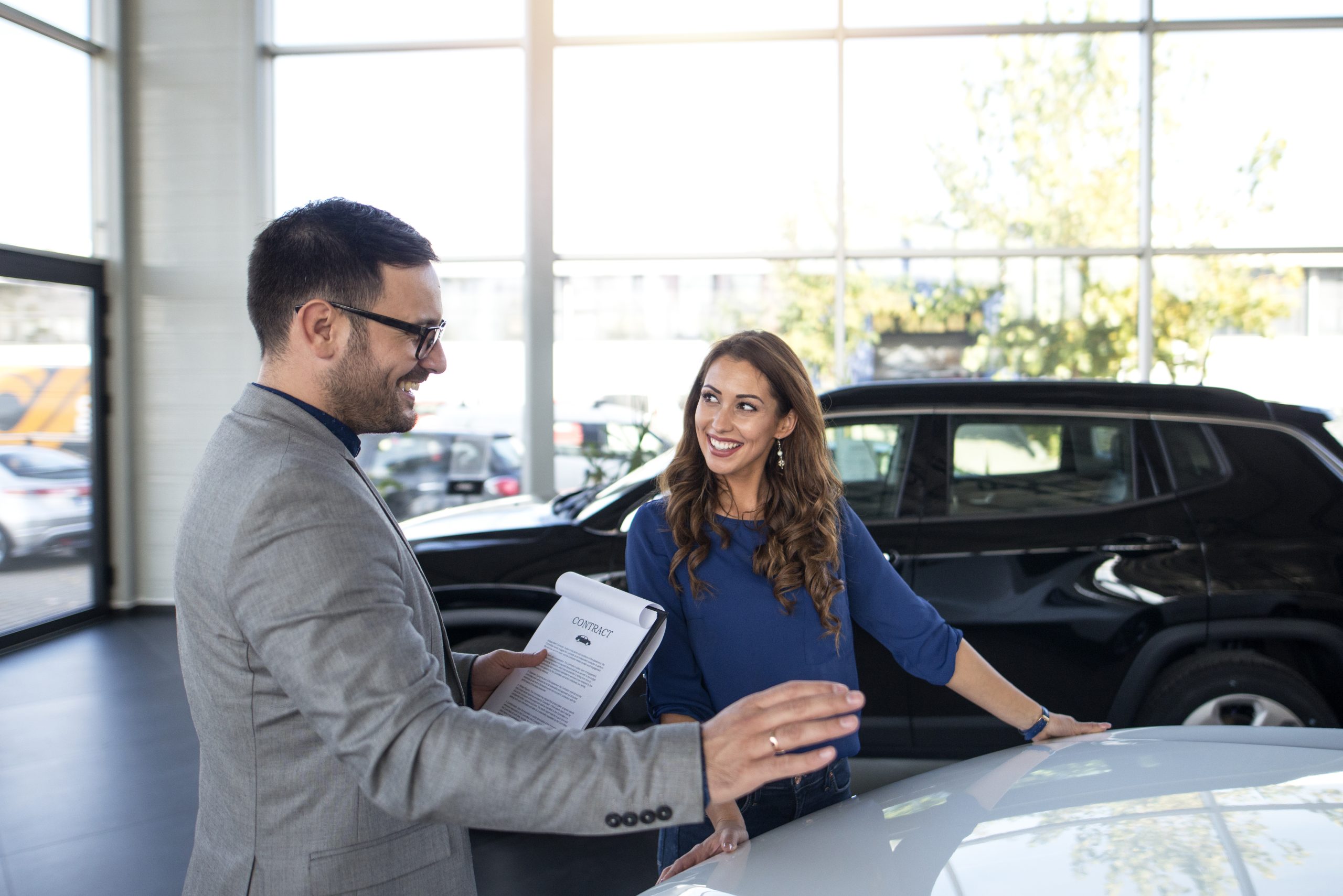 Una donna sorridente discute con un venditore all'interno di una concessionaria moderna e luminosa. Sullo sfondo, si intravedono diverse auto parcheggiate, mentre l'uomo, elegante con un completo grigio, tiene in mano un contratto. La scena rappresenta un momento positivo e cordiale, tipico del processo di finanziamento auto in Italia.