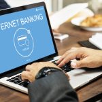 Un uomo in abito elegante utilizza un computer portatile su un tavolo di legno durante una colazione d'affari. Sullo schermo appare una schermata blu con la scritta "Internet Banking" e l'icona di una carta di credito con il saldo visibile. L'immagine rappresenta l'accesso digitale ai servizi bancari, un contesto che riflette le innovazioni introdotte da l'Open Banking, come la possibilità di gestire conti e transazioni in modo centralizzato e sicuro tramite applicazioni online.