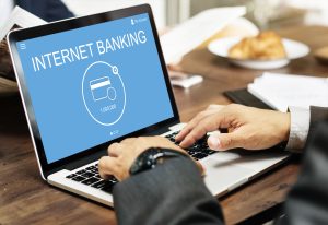 Un uomo in abito elegante utilizza un computer portatile su un tavolo di legno durante una colazione d'affari. Sullo schermo appare una schermata blu con la scritta "Internet Banking" e l'icona di una carta di credito con il saldo visibile. L'immagine rappresenta l'accesso digitale ai servizi bancari, un contesto che riflette le innovazioni introdotte da l'Open Banking, come la possibilità di gestire conti e transazioni in modo centralizzato e sicuro tramite applicazioni online.