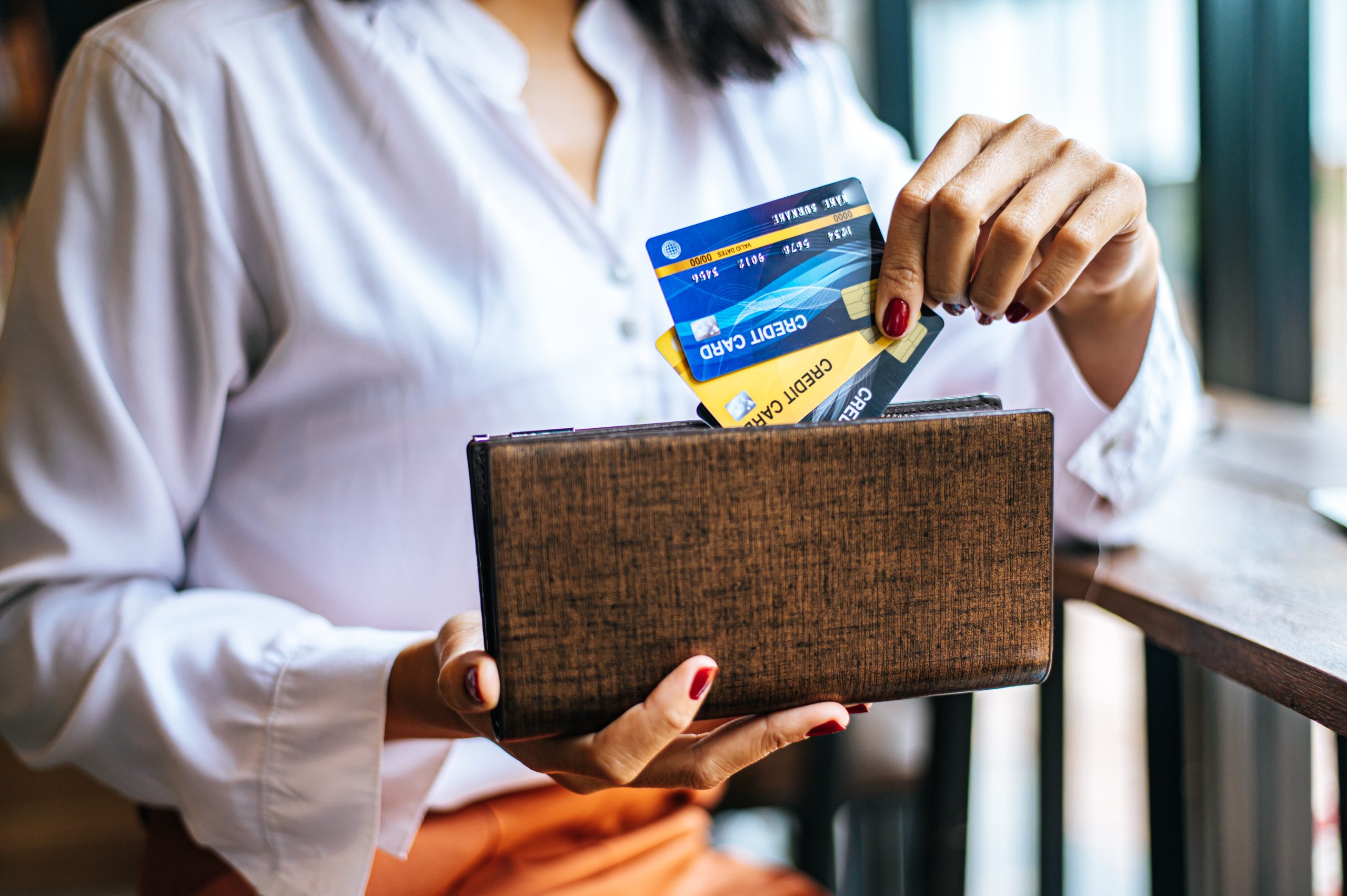 Donna seduta in un ambiente luminoso estrae diverse carte di credito da un portafoglio marrone. Indossa una camicia bianca elegante e ha le unghie laccate di rosso. L’immagine rappresenta una situazione tipica legata alla gestione finanziaria personale, perfetta per illustrare il concetto di trasferire il saldo tra carte di credito.