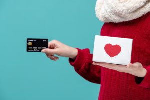 Persona in maglione rosso che porge una carta di credito nera mentre tiene in mano un cofanetto bianco con cuore rosso, simbolo di solidarietà e regalistica legata alle carte di credito con programma di donazione.