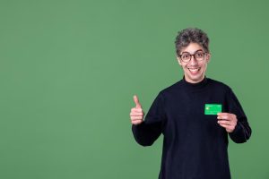 Uomo sorridente con occhiali e maglione scuro mostra il pollice in su mentre tiene una carta verde in mano, simbolo di approvazione e soddisfazione. Lo sfondo verde e l’espressione entusiasta trasmettono fiducia nei servizi bancari, ideali per promuovere carte di credito con benefici.