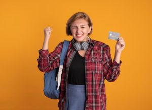 Studentessa giovane con zaino, cuffie al collo e camicia a quadri esprime gioia mentre mostra una carta di credito in mano, davanti a uno sfondo arancione acceso. L’immagine rappresenta perfettamente l’entusiasmo e le opportunità offerte dalle carte di credito per finanziare studi.