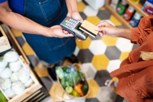 Una persona con una camicia arancione effettua un pagamento contactless con una carta su un terminale POS tenuto da un commesso con un grembiule blu, in un supermercato. In basso si intravede un cestino pieno di verdure fresche. Concetto di carte di credito per gli acquisti al supermercato.