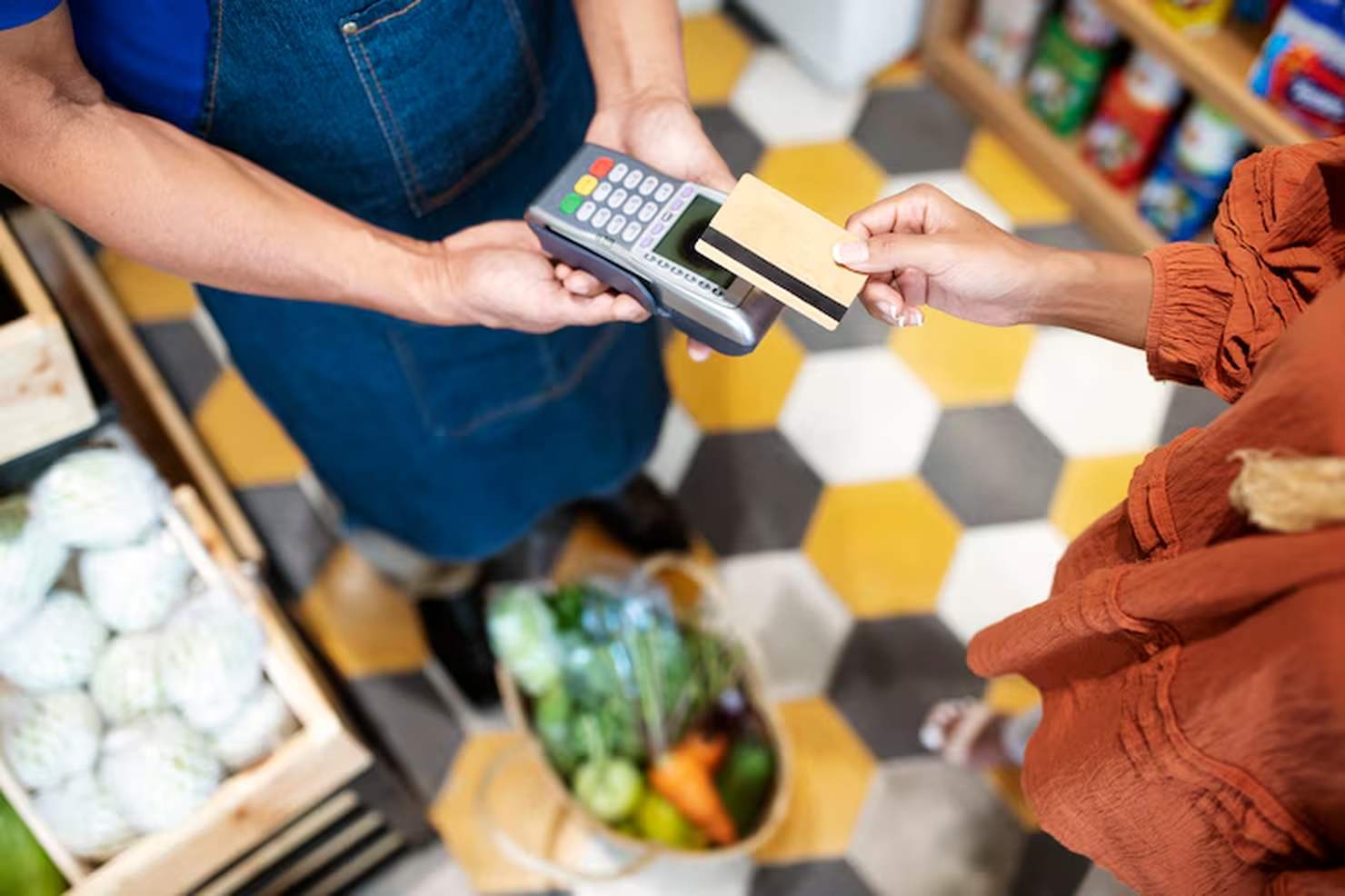Una persona con una camicia arancione effettua un pagamento contactless con una carta su un terminale POS tenuto da un commesso con un grembiule blu, in un supermercato. In basso si intravede un cestino pieno di verdure fresche. Concetto di carte di credito per gli acquisti al supermercato.