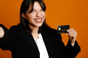 Donna sorridente con giacca nera che mostra con entusiasmo una delle sue carte di credito premium contro uno sfondo arancione vivace.