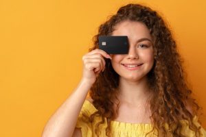 Giovane donna sorridente con capelli ricci tiene una carta nera davanti all’occhio, in posa su sfondo giallo, rappresentando l’uso moderno e pratico delle carte prepagate.