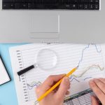Scrivania con laptop, calcolatrice, lente d’ingrandimento, grafici stampati e banconote, simboli di un’attenta pianificazione finanziaria. Le mani di una persona analizzano un foglio con dati percentuali e linee di tendenza. L’immagine rappresenta il monitoraggio dei costi fissi e variabili all’interno di un bilancio personale o aziendale, evidenziando l’importanza dell’organizzazione economica per una gestione efficace delle risorse.