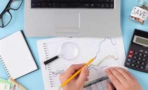 Scrivania con laptop, calcolatrice, lente d’ingrandimento, grafici stampati e banconote, simboli di un’attenta pianificazione finanziaria. Le mani di una persona analizzano un foglio con dati percentuali e linee di tendenza. L’immagine rappresenta il monitoraggio dei costi fissi e variabili all’interno di un bilancio personale o aziendale, evidenziando l’importanza dell’organizzazione economica per una gestione efficace delle risorse.