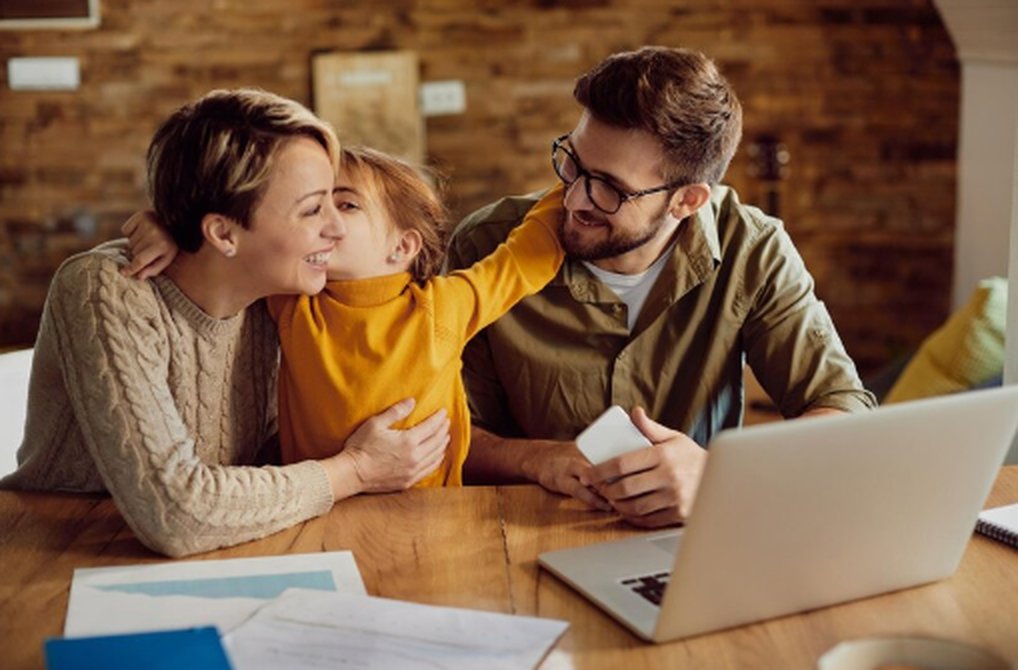 Famiglia seduta al tavolo con documenti e computer portatile, che sorride mentre discute insieme, rappresentando un momento di pianificazione finanziaria familiare in un ambiente domestico sereno.