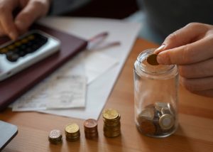 Una persona sta inserendo una moneta in un barattolo di vetro parzialmente pieno di spiccioli, mentre un’altra mano utilizza una calcolatrice accanto a documenti e ricevute su un tavolo. L'immagine rappresenta il concetto di risparmio personale e pianificazione finanziaria, con particolare attenzione alla creazione di una riserva di emergenza.