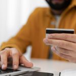 Un uomo, vestito con una camicia color senape, sta effettuando un pagamento online utilizzando una carta di credito mentre digita sul laptop. L’immagine rappresenta un acquisto digitale e richiama il concetto di sistema di carte a rate, evidenziando la praticità dei pagamenti dilazionati per la gestione delle spese personali.