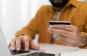 Un uomo, vestito con una camicia color senape, sta effettuando un pagamento online utilizzando una carta di credito mentre digita sul laptop. L’immagine rappresenta un acquisto digitale e richiama il concetto di sistema di carte a rate, evidenziando la praticità dei pagamenti dilazionati per la gestione delle spese personali.